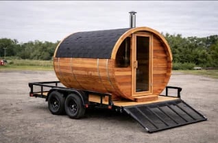 Wooden barrel-shaped mobile sauna trailer with ramp parked on asphalt surface
